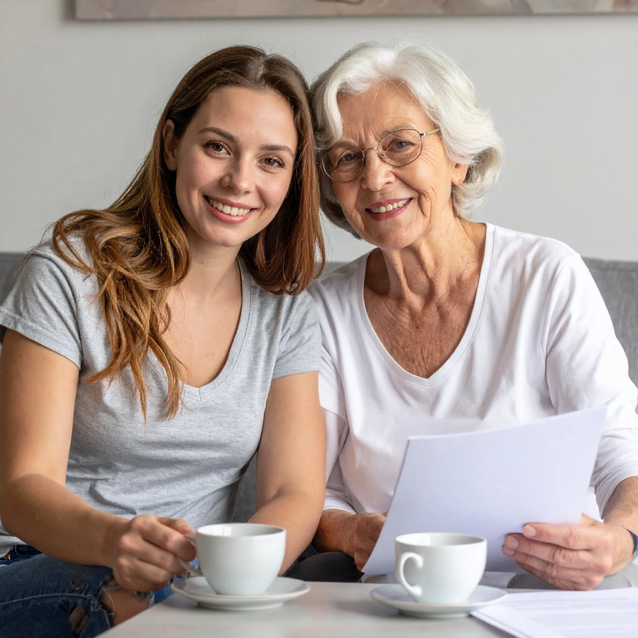 Two women sitting at a table with two white coffee cups, smiling, having a conversation, with one holding a paper.