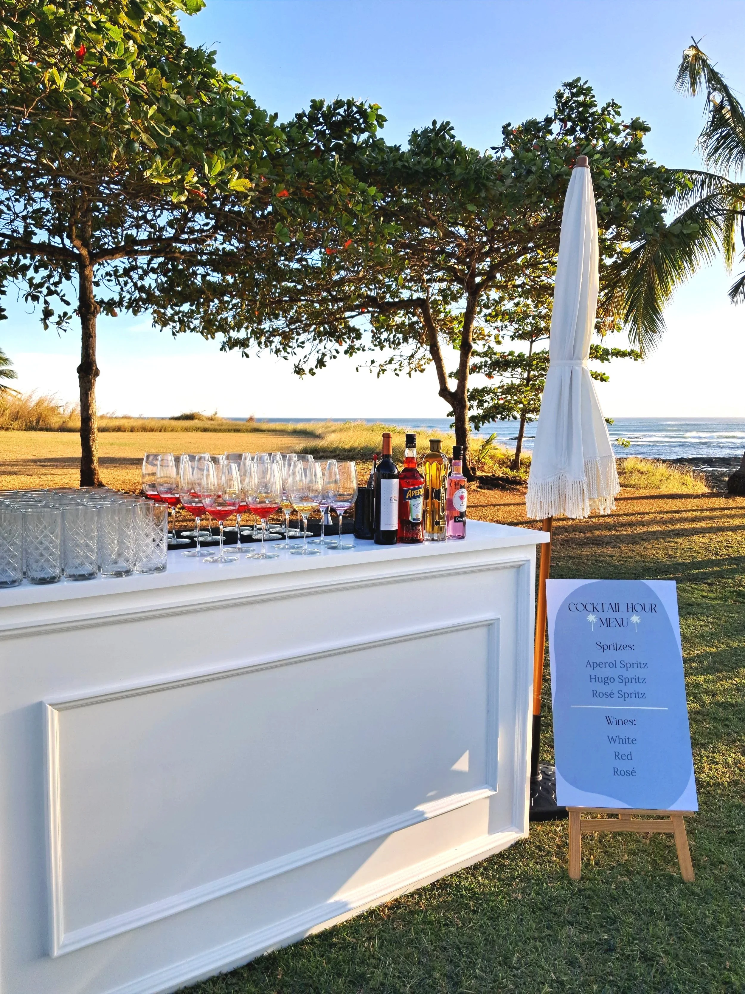 Outdoor bar setup with wine glasses, liquor bottles, and a cocktail menu on a signboard next to a white bar counter at the beach, with trees, palm trees, and ocean in the background.