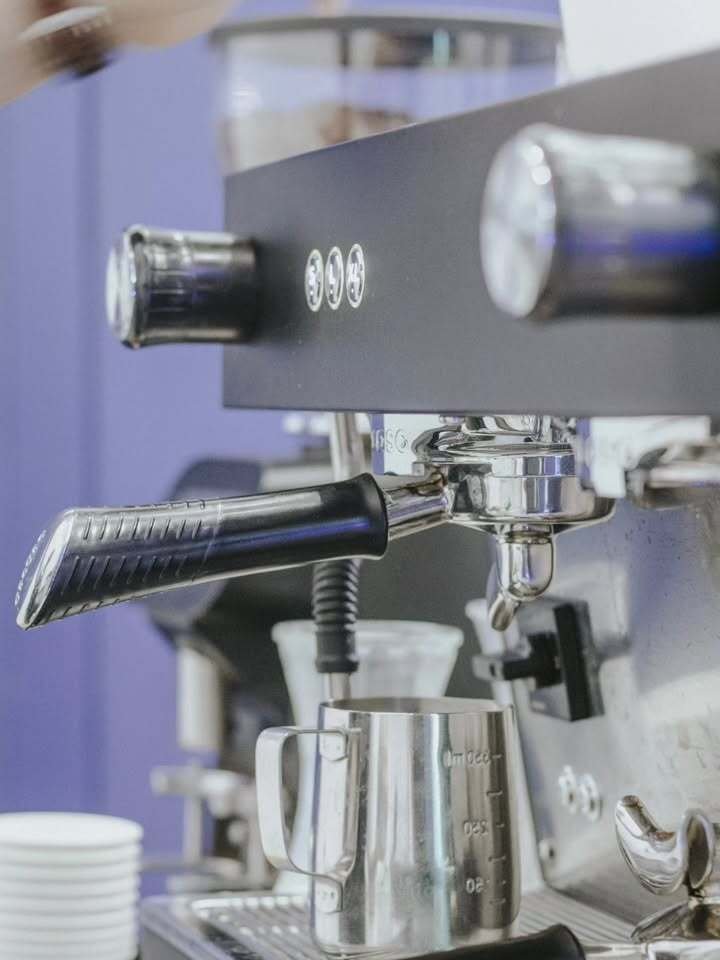 A close-up of a professional espresso machine with a portafilter inserted, a stainless steel milk frothing pitcher underneath, and a few control knobs and indicator lights on the machine.