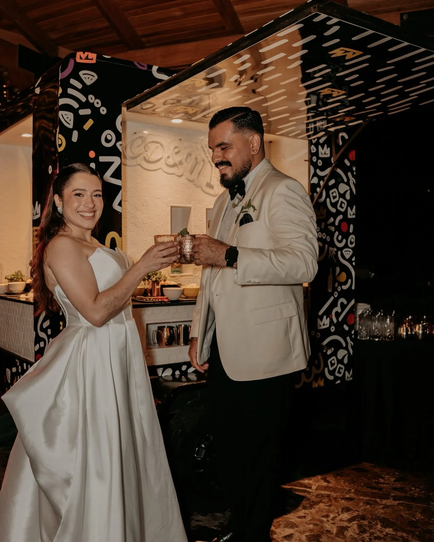 A bride and groom sharing a toast at their wedding reception, smiling and dressed in formal attire. The bride is wearing a white wedding gown and the groom is in a cream-colored tuxedo with a black bowtie. They are indoors with a decorated background and a bar area.