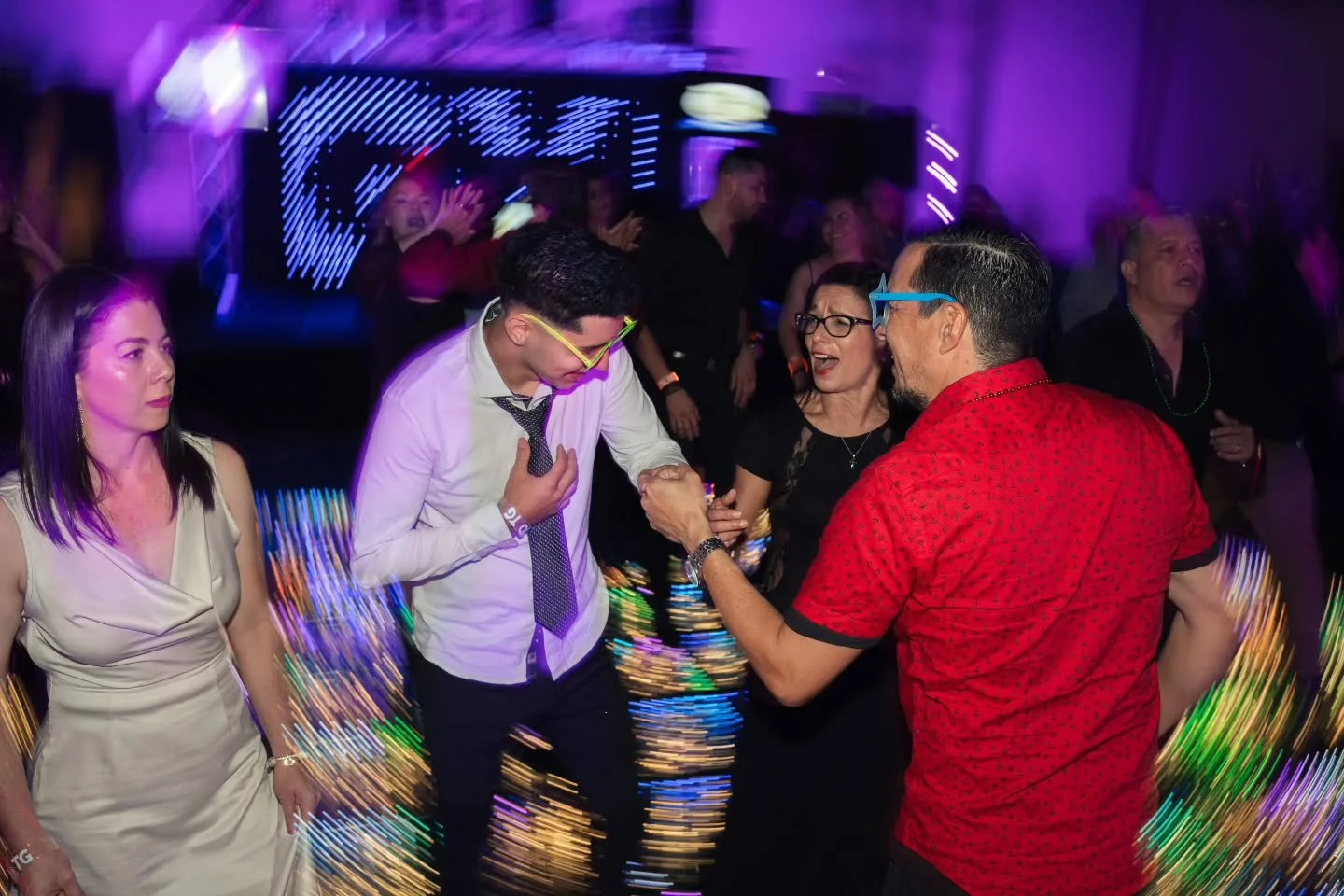 People dancing and socializing at a nightclub with colorful lights and a purple hue, including a man in a white shirt and tie, a woman in black, and a man in a red shirt with glasses, with some motion blur and light streaks.