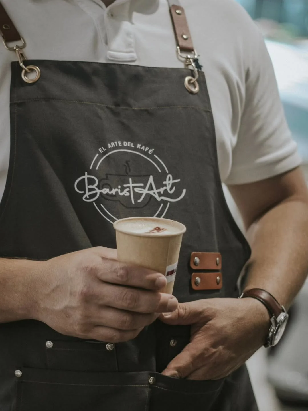 Person wearing a black apron with the logo 'BaristArt' holding a cup of coffee.