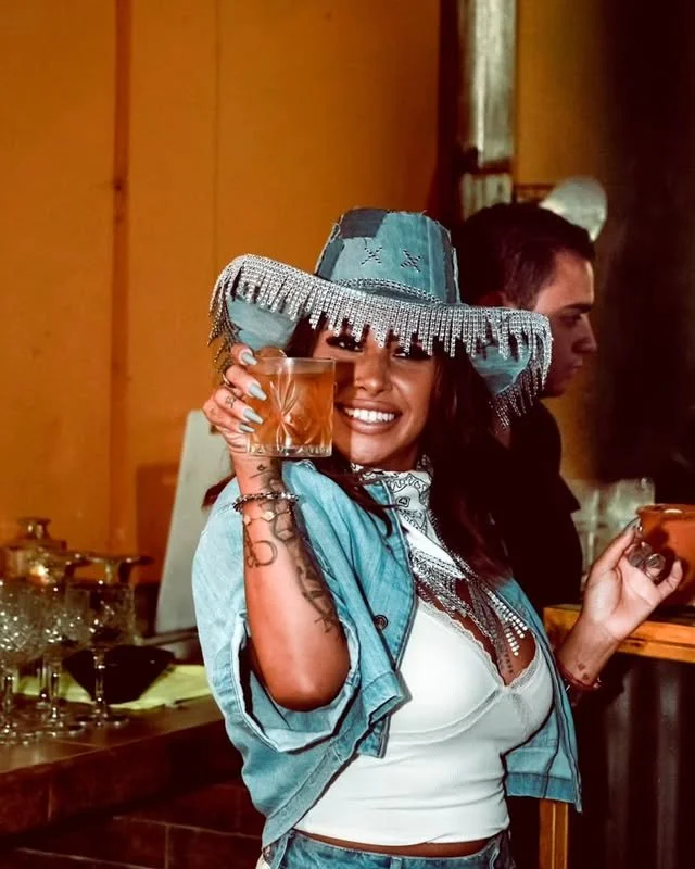 A woman wearing a festive sombrero hat, smiling and holding a glass of amber drink at a celebration or party.
