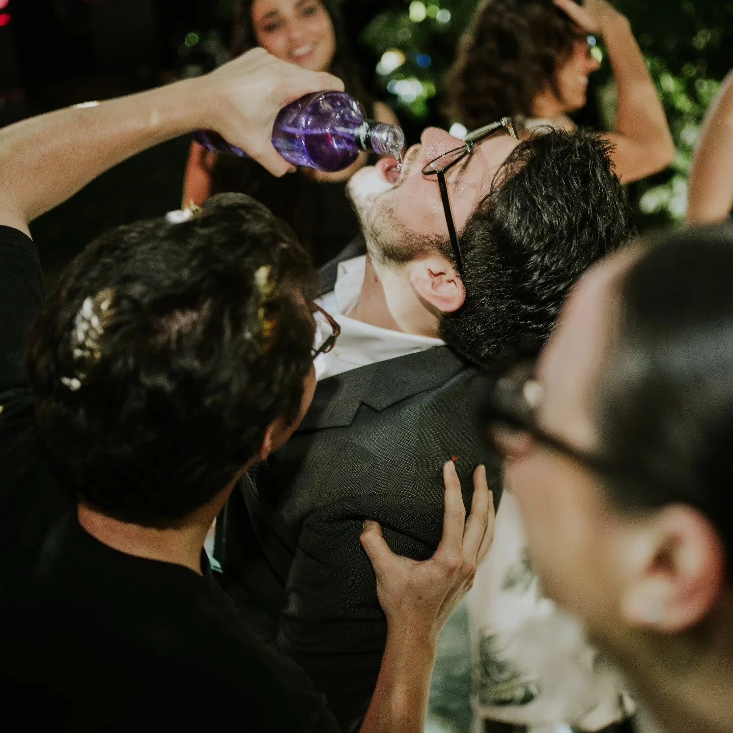 A man with glasses and a beard lying down while a woman pours a drink into his mouth at a social gathering.