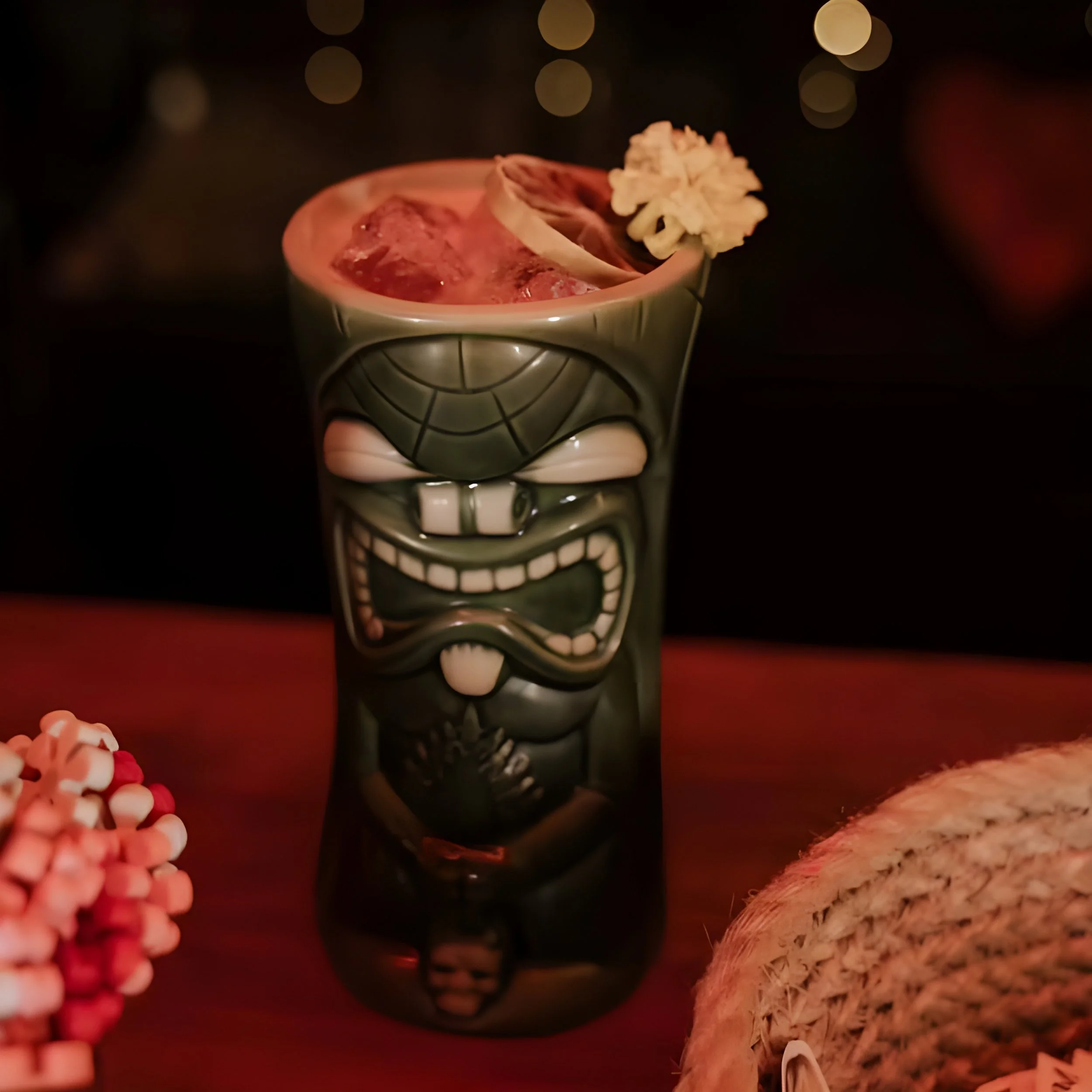 A close-up photo of a decorative cup with a skeleton face design, featuring a wide, toothy smile, holding a drink with ice and a slice of fruit, with a dark background and blurred lights.