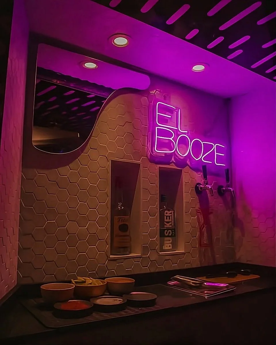 Bar area with hexagonal tile wall, neon purple sign that reads 'EL BOOZE', and skateboards mounted on the wall, illuminated with purple lighting.