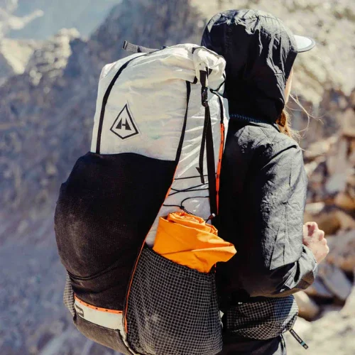 Person wearing a black hooded jacket and cap, carrying a large Hyperlite hiking backpack in a rocky outdoor area.