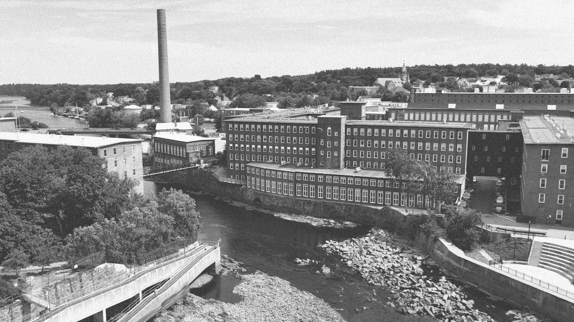 Black and white aerial view of the Pepperell Mill campus with buildings, a river, and a smokestack in the background.