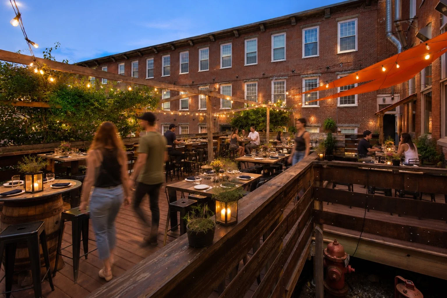Outdoor rooftop dining area at dusk with string lights, tables with candles and flowers, and people enjoying their evening in front of brick buildings.