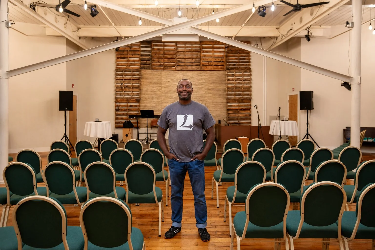 A man standing in an empty conference or event room with green chairs arranged in rows, a stage with musical equipment and microphones at the back, and string lights hanging from the ceiling.
