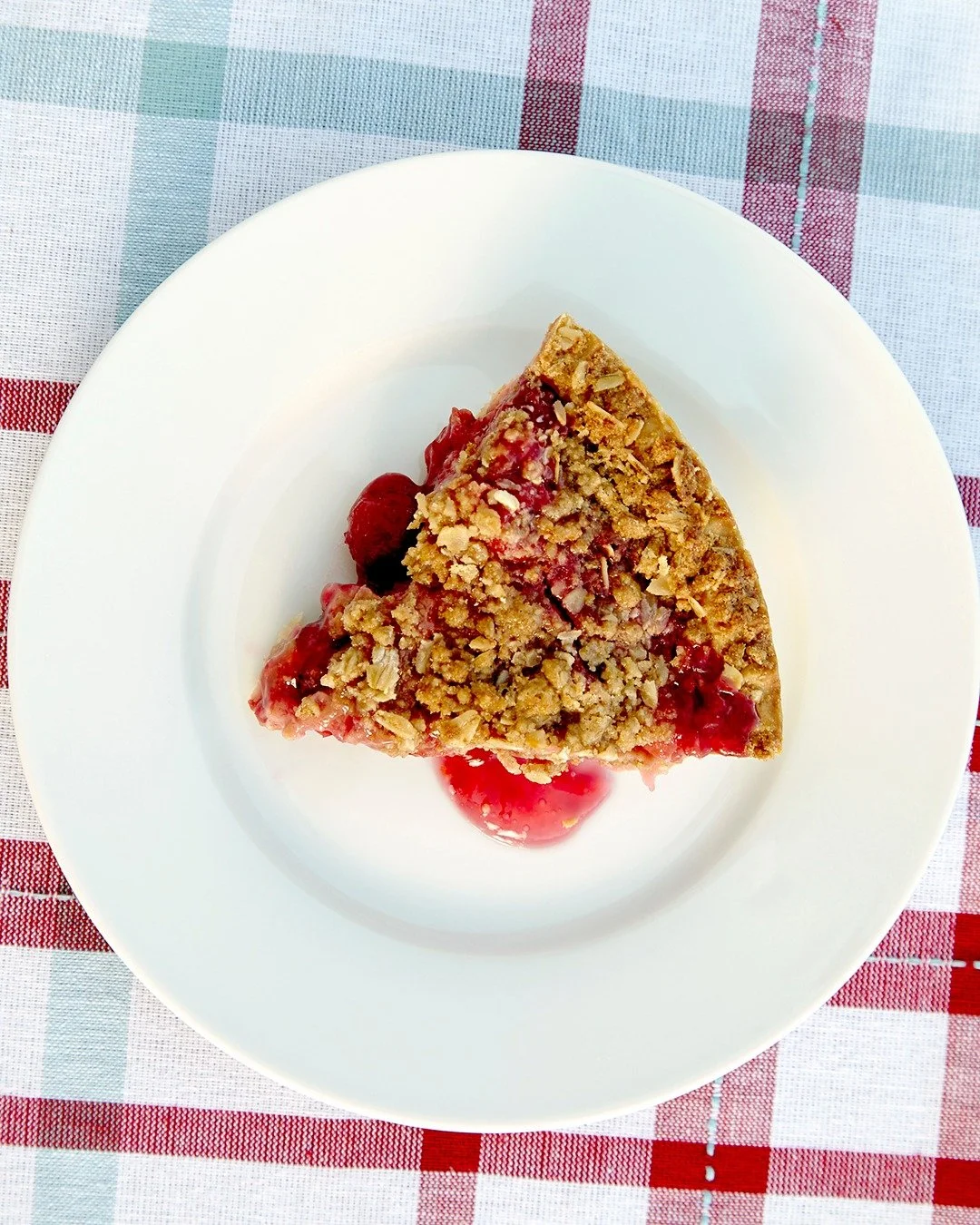 A slice of cherry crumble pie from Maine Pie Co. on a white plate with a plaid tablecloth in the background.