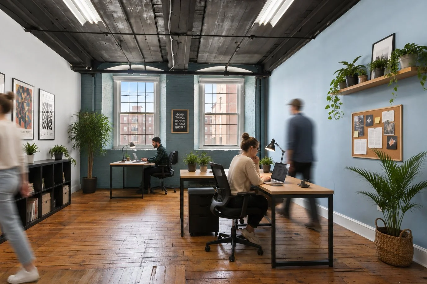 A modern office with wooden floors, white and light blue walls, and large windows. Desks with laptops and plants. People working, one woman at a desk and two men walking. Wall art and cork board decor. Potted plants on shelves and around the room.