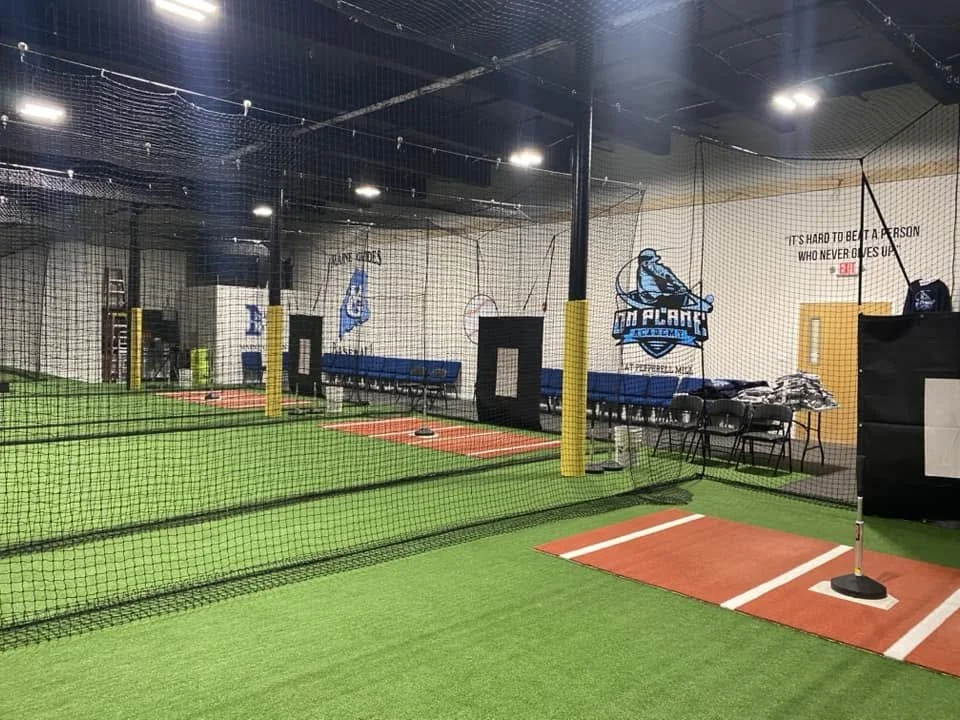 Indoor baseball or softball batting practice area with artificial turf, nets, and a batting tee.