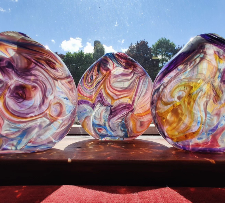 Three abstract swirled glass art pieces with vibrant colors, displayed on a wooden surface with a sunny outdoor background and blue sky.