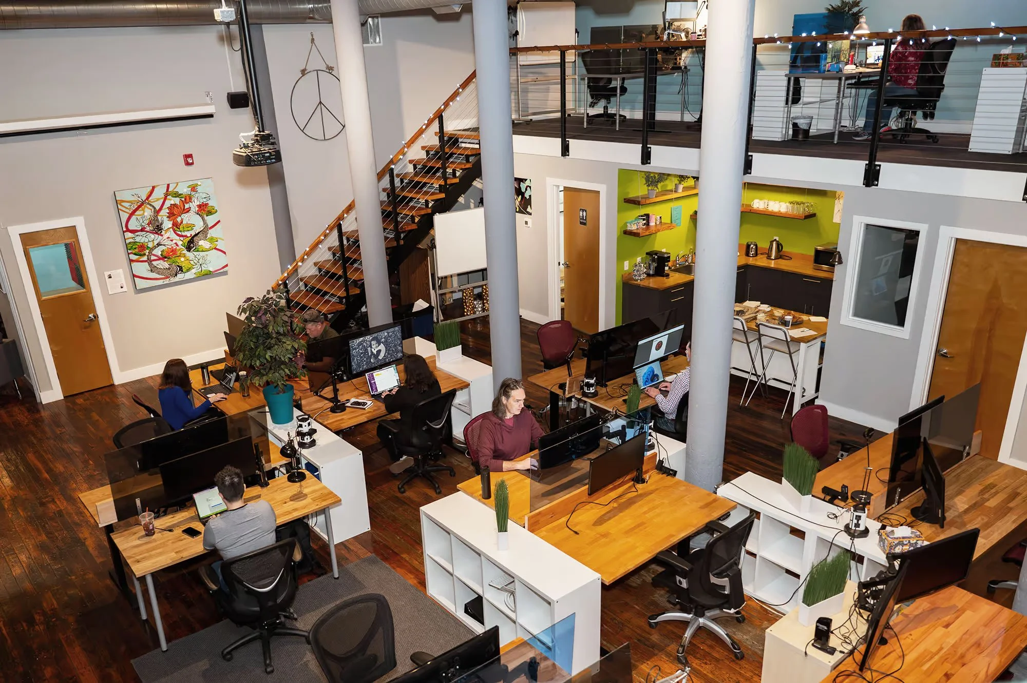 Co-working space with multiple people working at desks with computers, a staircase leading to an upper level with more workspaces, and decorative elements like colorful artwork and plants.