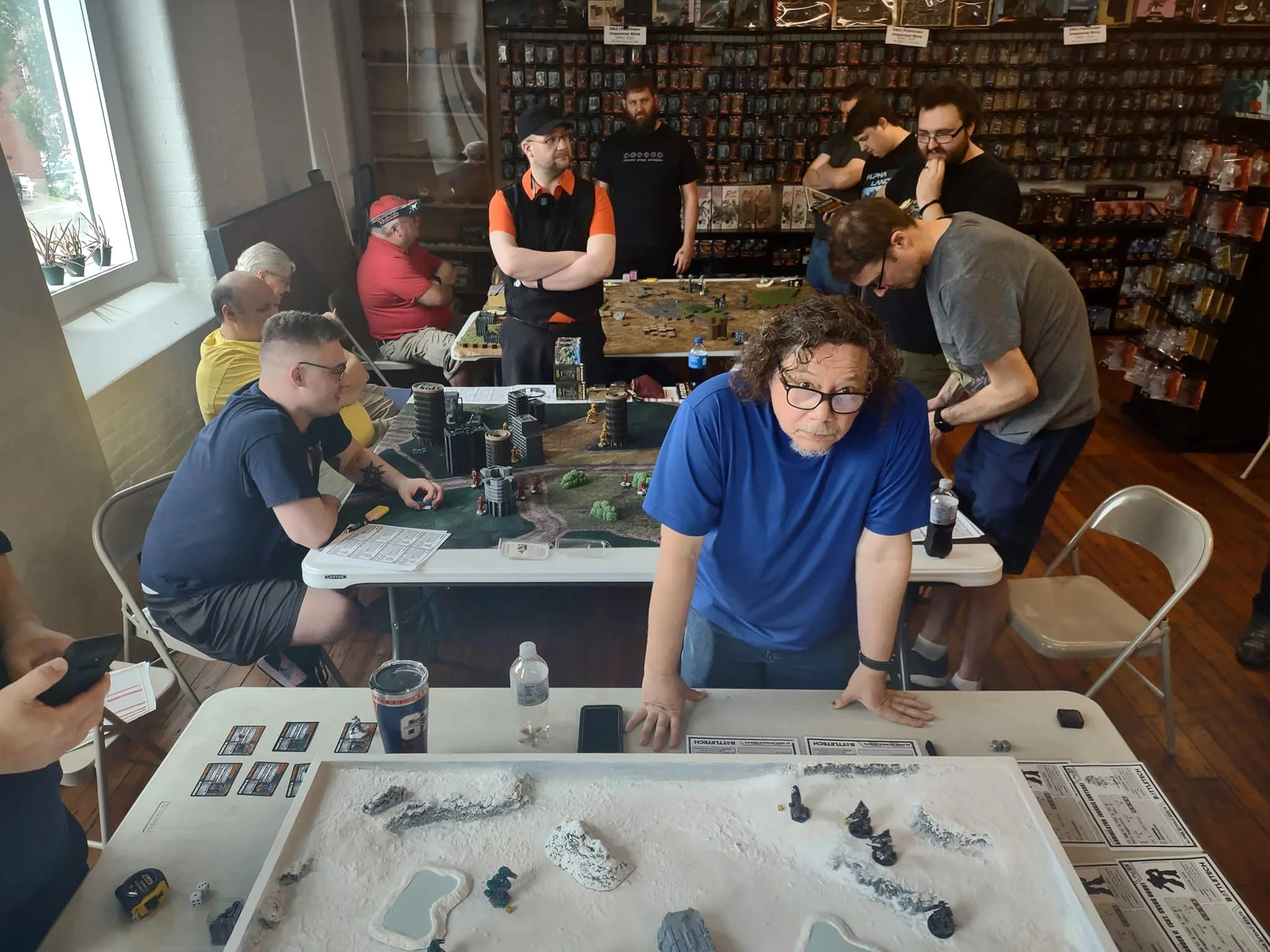 Group of people playing a tabletop miniature game in a store with shelves of miniature figurines on the wall. The table has miniatures, dice, and terrain pieces, with some participants focused on gameplay and others observing.