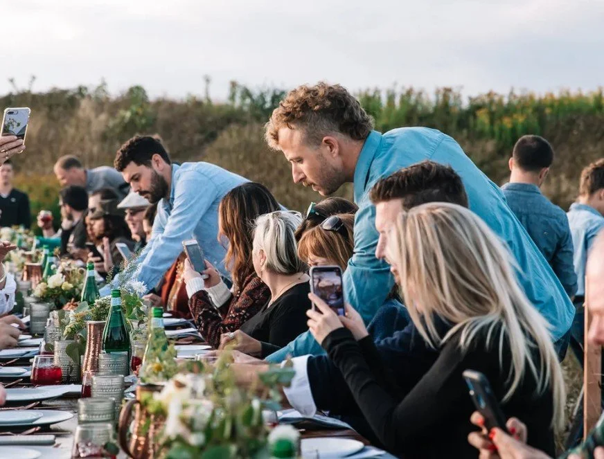 People gathered around a long outdoor dining table, taking photos and enjoying a meal during a social event.