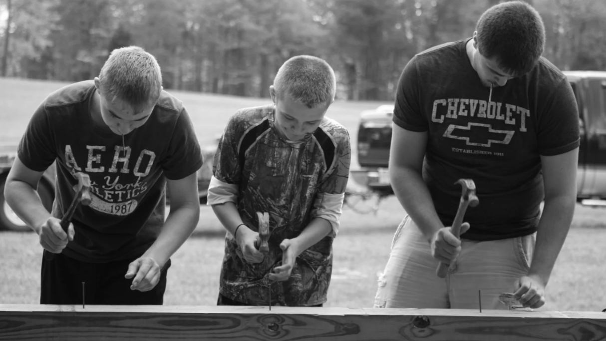Three boys working with hammers on a wooden project outdoors with trees and parked vehicles in the background.