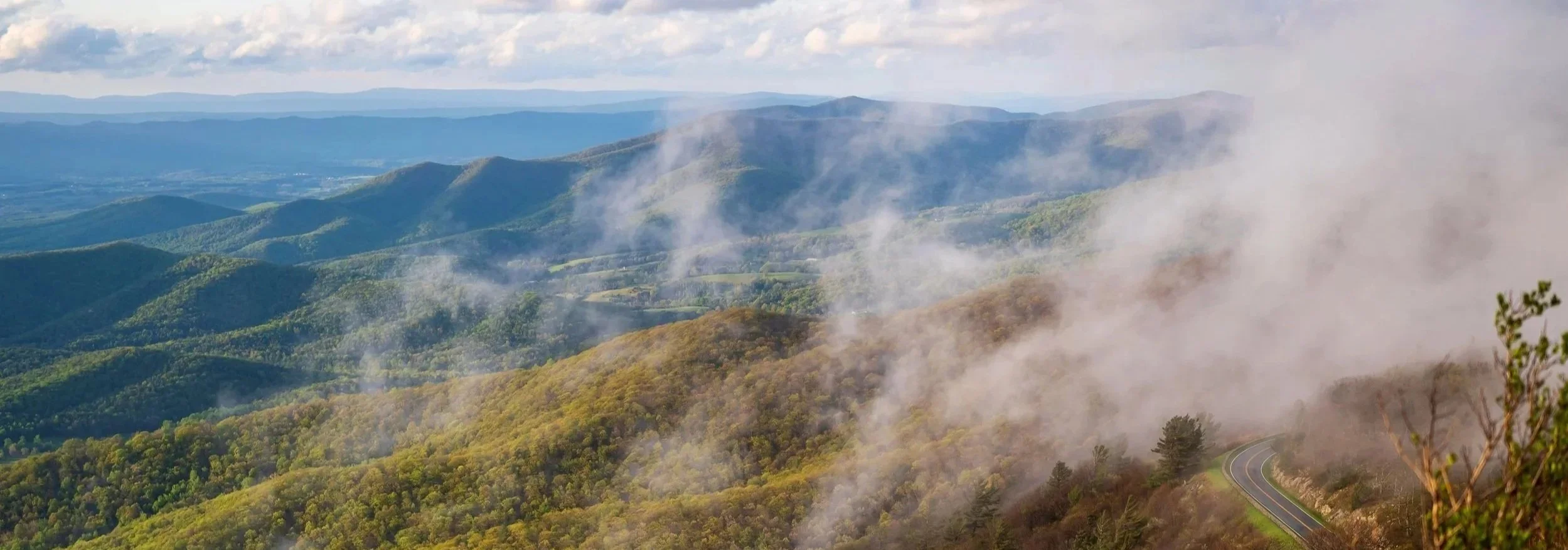 A view of green mountain ranges with smoke or fog rising among the trees, a winding road visible at the bottom right corner, under a partly cloudy sky.