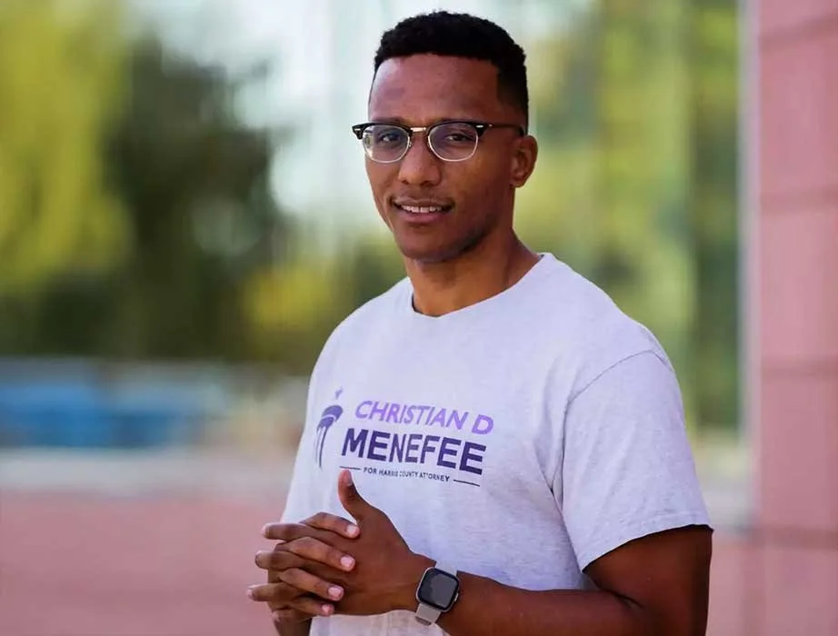 Young man wearing glasses and a gray T-shirt standing outdoors with blurred greenery background.