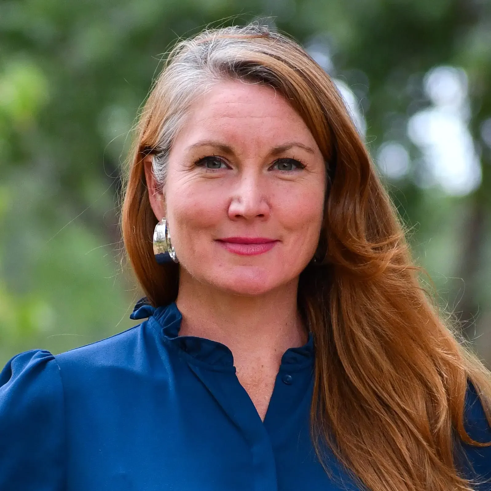 A woman with long red hair and silver hoop earrings outdoors. She is wearing a blue blouse with a ruffled collar and smiling slightly. The background is blurred with green foliage.