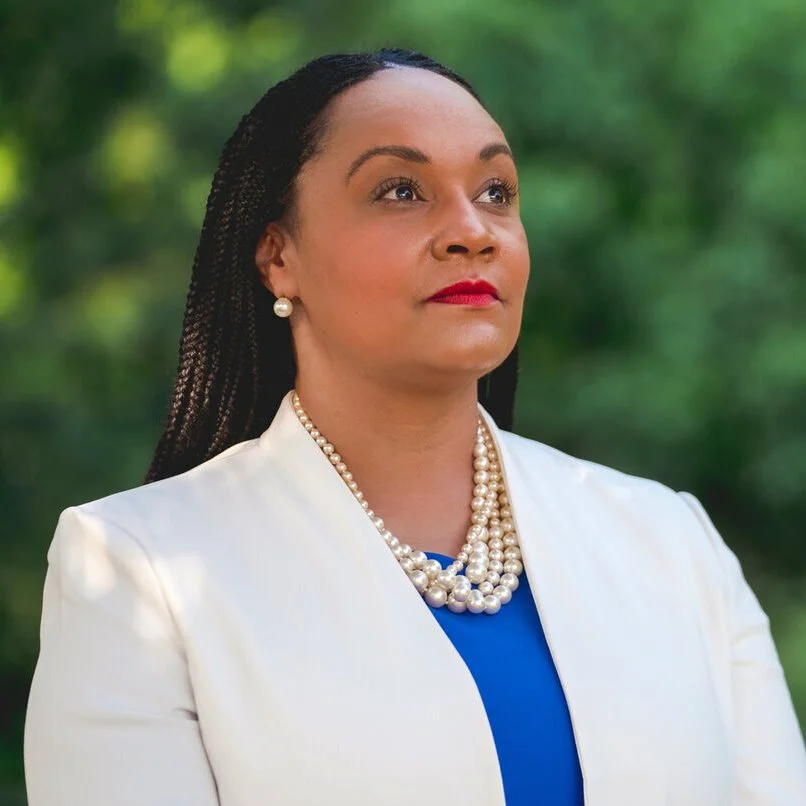 A woman with braided hair, wearing pearl earrings and a multi-strand pearl necklace, dressed in a white blazer over a blue top, standing outdoors with a blurred green background.