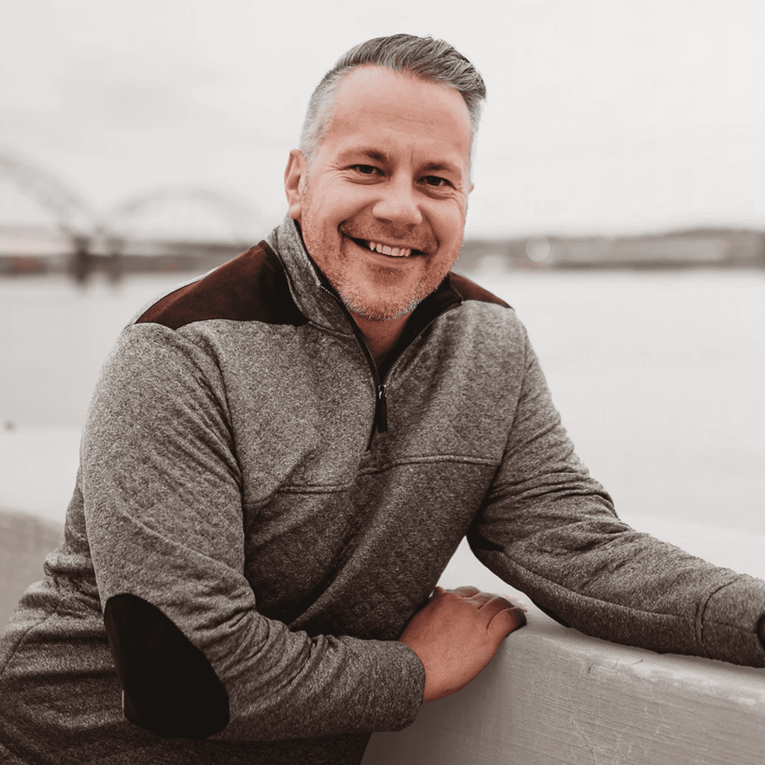A smiling man with gray hair and a beard, wearing a gray jacket with black accents, leaning on a white fence in front of a body of water and bridge in the background.