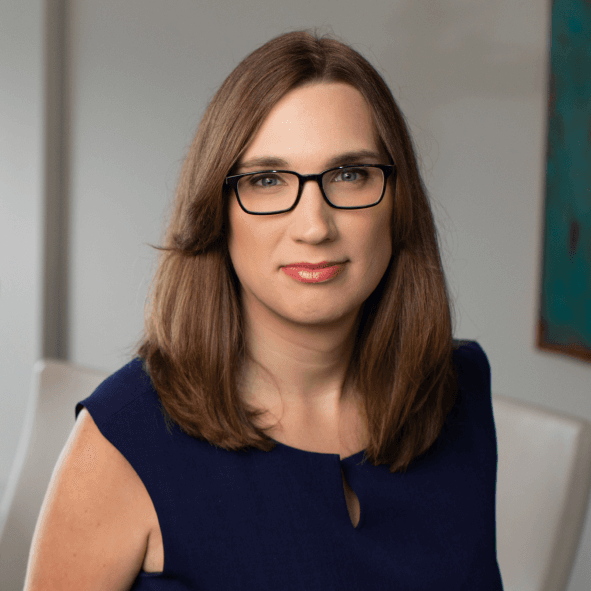 A woman with shoulder-length brown hair, wearing glasses and a navy blue sleeveless top, sitting in a room with modern decor.