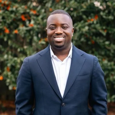 A man in a dark blue suit and white shirt smiling outdoors with greenery and orange flowers in the background.