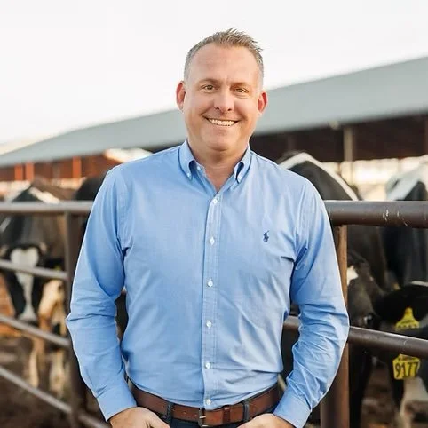 A man smiling outdoors in front of a farm with black and white cows.