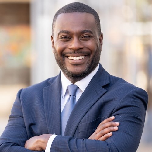 A smiling Black man in a blue suit with crossed arms.