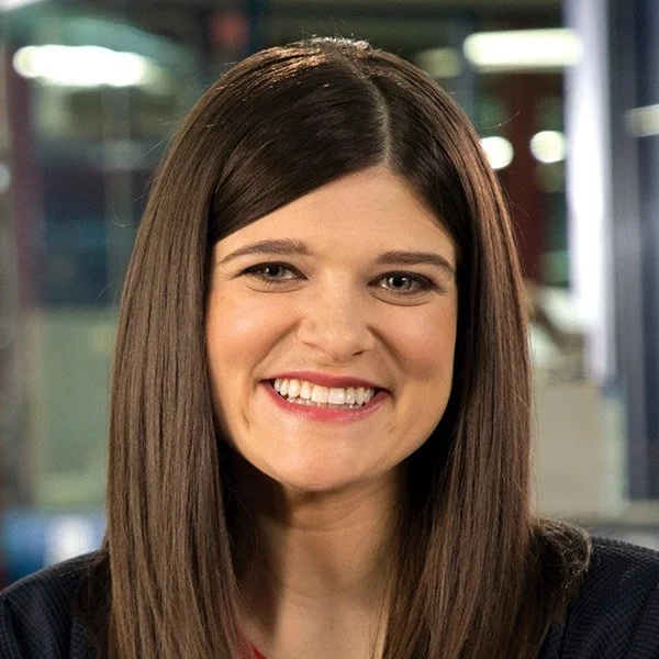 Close-up of a smiling woman with straight brown hair, wearing a dark top, in an indoor setting with blurred background.