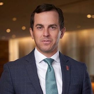 A man in a dark suit with a white shirt and teal tie, standing indoors with blurred warm lighting in the background.