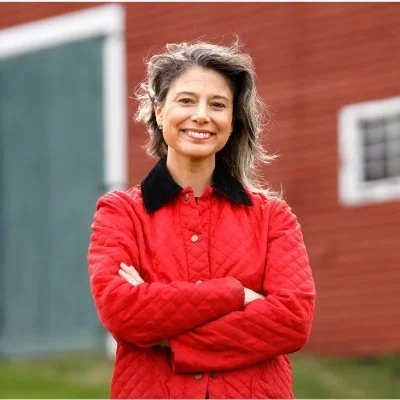 A woman with gray hair smiling with arms crossed, wearing a red quilted jacket with black collar, standing outdoors in front of a red building and green chalkboard.