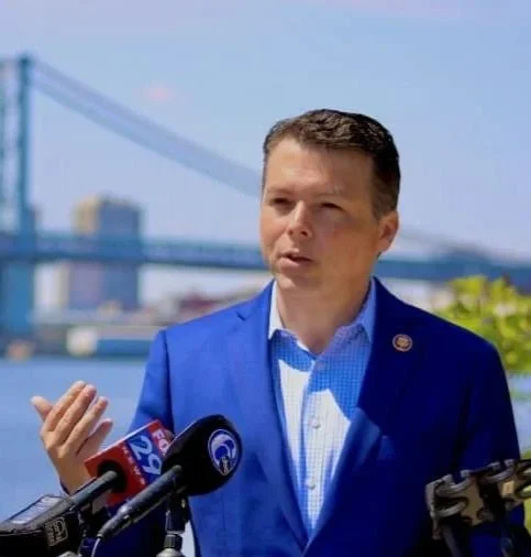 A man in a blue suit speaking at a press conference outdoors, with a bridge and cityscape in the background.