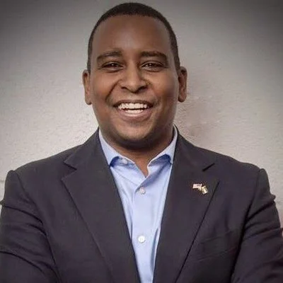 A smiling man in a suit with a small American flag pin on his lapel, posing against a neutral background.