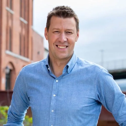 A smiling man in a blue button-up shirt standing outdoors in front of buildings and a cloudy sky.