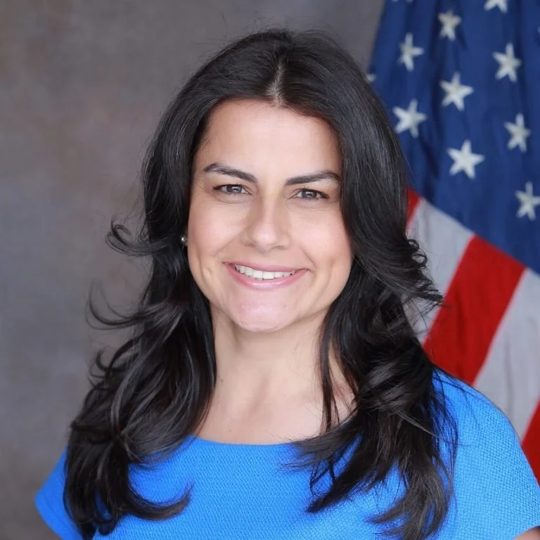 A woman with long dark hair wearing a blue top standing in front of the American flag.