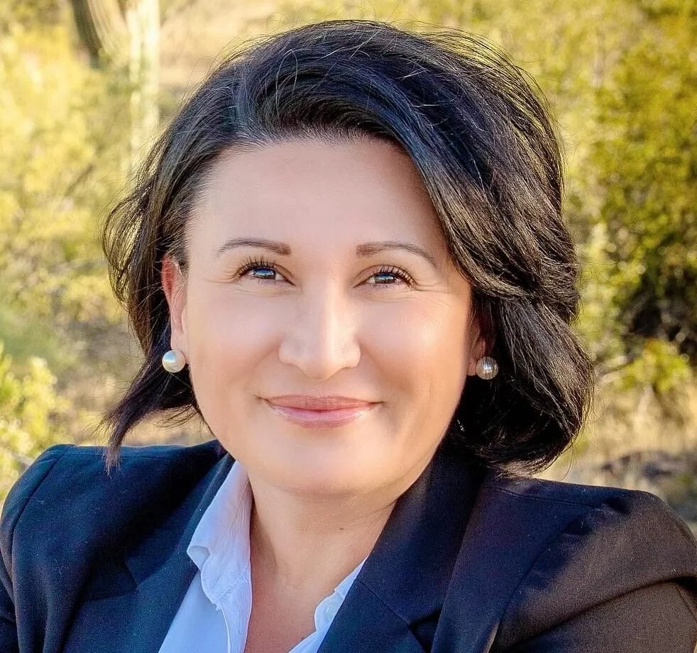 Headshot of a smiling woman with short dark hair, wearing pearl earrings and a dark blazer, outdoors with greenery in the background.