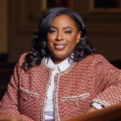 A smiling woman with dark curly hair wearing a red textured blazer over a white shirt, seated indoors with a wooden background.