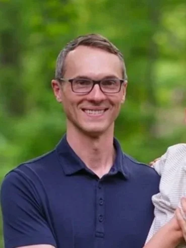 A man with glasses smiling outdoors on a green, blurred background.