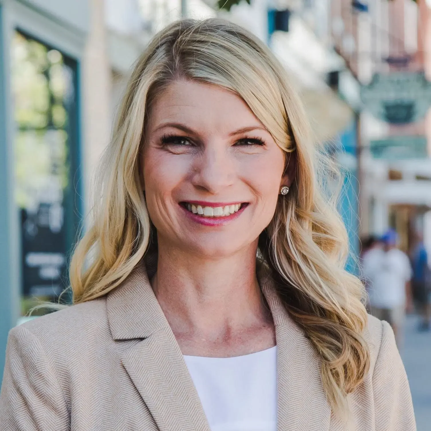 A smiling woman with blonde hair wearing a beige blazer and white top, standing outdoors in an urban area with buildings and blurred people in the background.