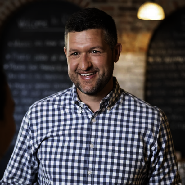 A man smiling, wearing a blue and white checkered shirt, in a cozy cafe or restaurant setting with warm lighting and a chalkboard menu in the background.
