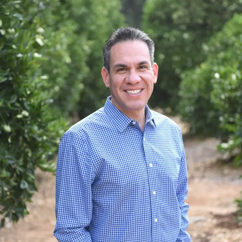 Smiling man in blue checkered shirt outdoors among green trees.