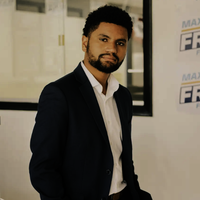 A man with curly black hair and a beard wearing a navy blazer and white shirt, standing indoors next to a window with a white wall and a banner in the background.