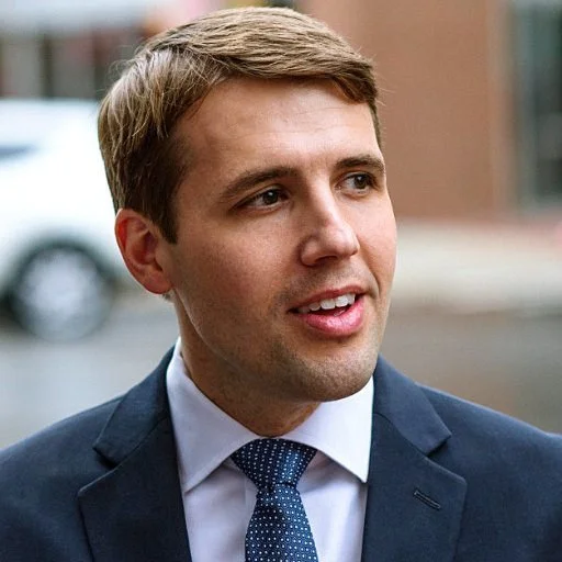 A young man in a navy suit and blue tie standing outdoors, speaking or listening, with a slight smile.