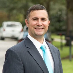 A man in a dark suit with a light blue tie, smiling outdoors with cars and trees in the background.