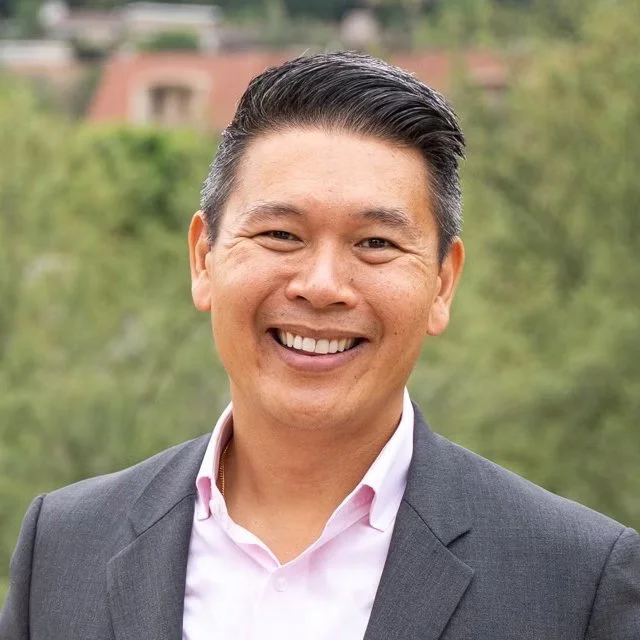 A smiling man in a gray suit and pink dress shirt outdoors with blurred trees and buildings in the background.