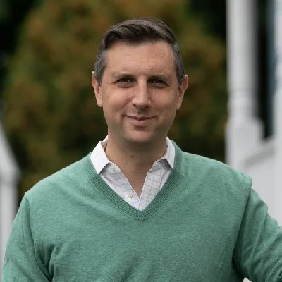 A man with short dark hair wearing a green V-neck sweater over a collared shirt, standing outdoors with trees in the background.