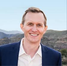 A man with short hair in a suit smiling outdoors with mountains in the background.